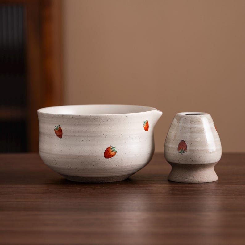 May include: A ceramic matcha bowl and whisk holder set. The bowl is off-white with a spout and small red strawberry designs. The whisk holder is a matching off-white color with a strawberry design. The set is designed for preparing matcha tea.