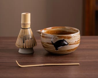 Op de afbeelding: Een bruine en witte keramische matcha-kom met een zwart kattenontwerp. De kom heeft een tuit en staat op een houten tafel. Een bamboe matcha-garde en een bamboe matcha-schep staan ook op de tafel.