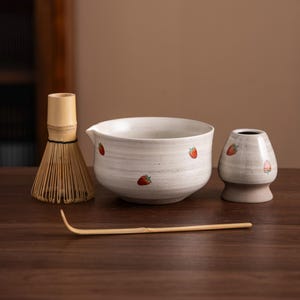 May include: A matcha tea set featuring a bamboo whisk, a ceramic bowl with a spout, a ceramic tea caddy, and a bamboo scoop. The bowl and caddy are off-white with red strawberry designs. The set is arranged on a wooden surface.