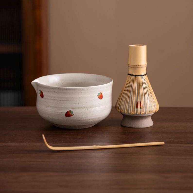 May include: A matcha tea set featuring a ceramic bowl with strawberry designs, a bamboo whisk, and a bamboo scoop. The bowl is light gray with red strawberry illustrations. The whisk is made of bamboo and has a light brown color. The scoop is also made of bamboo.