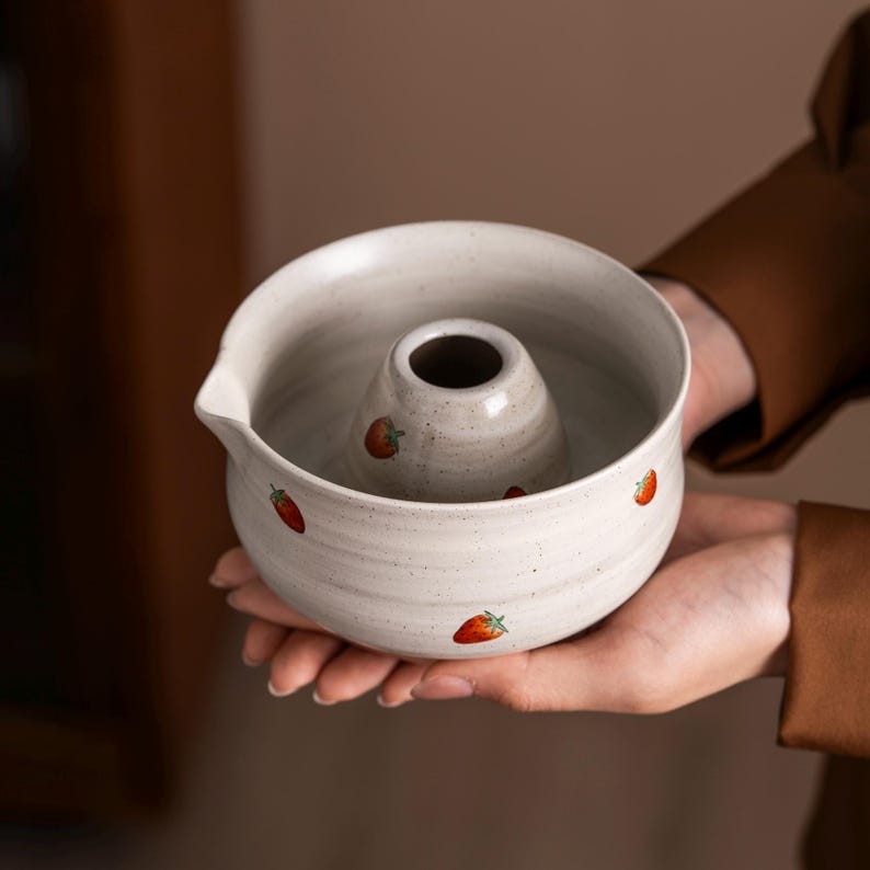 May include: A speckled white ceramic bundt pan with a central cone and a pouring spout. The pan is decorated with small red strawberry illustrations. The pan is held in a person's hands.