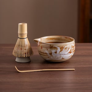 Puede incluir: Un cuenco de matcha de cerámica marrón y blanco con un diseño de fénix blanco, un batidor de matcha de bambú y una cuchara de matcha de bambú sobre una superficie de madera.