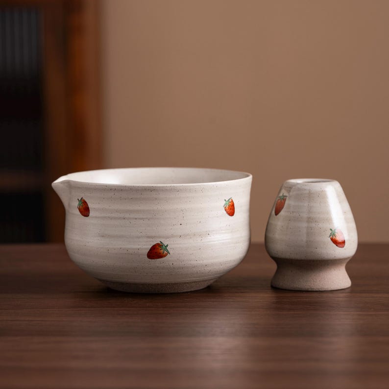 May include: A ceramic tea bowl and whisk holder set. The off-white pieces are decorated with red strawberry illustrations. The bowl has a spout. The whisk holder has a rounded shape and a textured base.