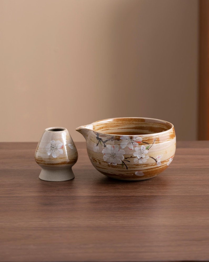 May include: A set of two ceramic teacups with a brown and white striped pattern and white cherry blossom designs. The smaller cup has a rounded shape and the larger cup has a spout.
