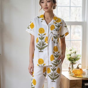May include: A white pajama set featuring a short-sleeved button-down shirt and matching pants, both decorated with a repeating pattern of yellow marigold flowers and green stems. The shirt includes a notched collar and a single chest pocket.
