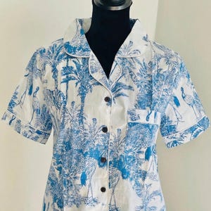 Op de afbeelding: Wit overhemd met een blauwe tropische print met palmbomen en vogels. Het shirt met korte mouwen heeft een reverskraag, een borstzak en donkerbruine knopen. Het shirt maakt deel uit van een pyjamaset.