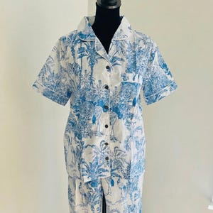 Op de afbeelding: Witte en blauwe pyjamaset met tropische print. De set bestaat uit een shirt met korte mouwen en knopen en een bijpassende broek. De print toont palmbomen, vogels en andere planten. Het shirt heeft een borstzak en witte bies.