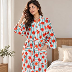 May include: A long-sleeved robe featuring a repeating pattern of bright orange sunbursts over vertical light blue and white stripes. The garment includes a matching fabric tie belt at the waist and two front patch pockets on the lower section.