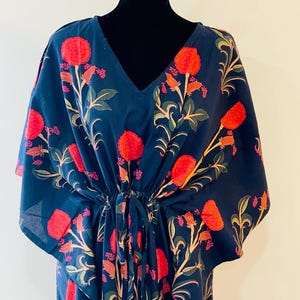 K&ouml;nnte beinhalten: Ein marineblaues Kaftankleid mit V-Ausschnitt und Kordelzug in der Taille. Das Kleid hat ein Blumenmuster mit roten Bl&uuml;ten, gr&uuml;nen Bl&auml;ttern und beigen Stielen. Der Kaftan wird auf einer schwarzen Schaufensterpuppe pr&auml;sentiert.
