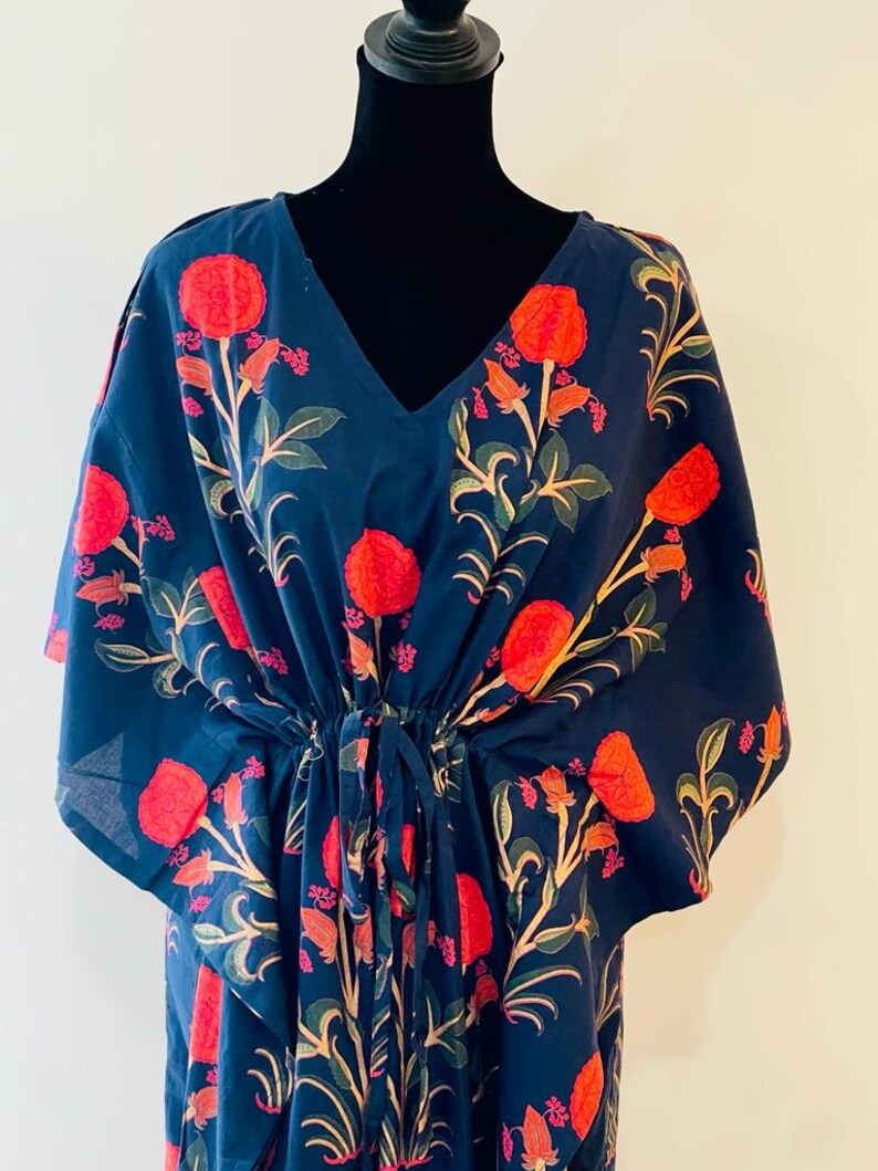 K&ouml;nnte beinhalten: Ein marineblaues Kaftankleid mit V-Ausschnitt und Kordelzug in der Taille. Das Kleid hat ein Blumenmuster mit roten Bl&uuml;ten, gr&uuml;nen Bl&auml;ttern und beigen Stielen. Der Kaftan wird auf einer schwarzen Schaufensterpuppe pr&auml;sentiert.