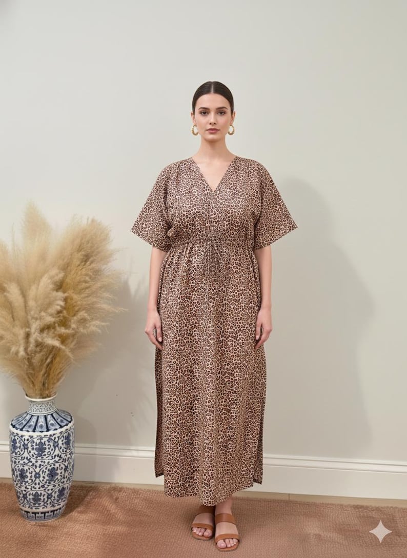 K&ouml;nnte beinhalten: Ein langes, braunes Kleid mit Leopardenmuster, kurzen &Auml;rmeln und V-Ausschnitt. Das Kleid hat eine Kordel in der Taille und Seitenschlitze. Das Model tr&auml;gt goldene Creolen und braune Sandalen. Im Hintergrund eine Vase mit hohen, getrockneten Pflanzen.
