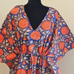K&ouml;nnte beinhalten: Ein blauer Kaftan mit einem leuchtenden Blumenmuster in Orange und Wei&szlig;. Der V-Ausschnitt-Kaftan ist aus leichtem Stoff gefertigt und hat eine Kordel in der Taille. Das florale Design zeigt gro&szlig;e, stilisierte Blumen und Bl&auml;tter.