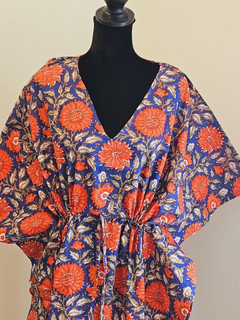 K&ouml;nnte beinhalten: Ein blauer Kaftan mit einem leuchtenden Blumenmuster in Orange und Wei&szlig;. Der V-Ausschnitt-Kaftan ist aus leichtem Stoff gefertigt und hat eine Kordel in der Taille. Das florale Design zeigt gro&szlig;e, stilisierte Blumen und Bl&auml;tter.