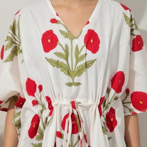 K&ouml;nnte beinhalten: Wei&szlig;es Kaftan-Kleid mit rotem Blumendruck. Das Kleid hat eine Taille mit Kordelzug und kurze &Auml;rmel. Das florale Design umfasst rote Mohnblumen und gr&uuml;ne Bl&auml;tter.