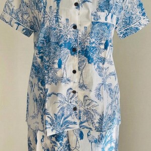 Op de afbeelding: Wit en blauwe pyjamaset met een tropische toile de Jouy-print met palmbomen, zebra's en vogels. De top met korte mouwen heeft een knoopsluiting en een borstzak. De bijpassende broek maakt de set compleet.