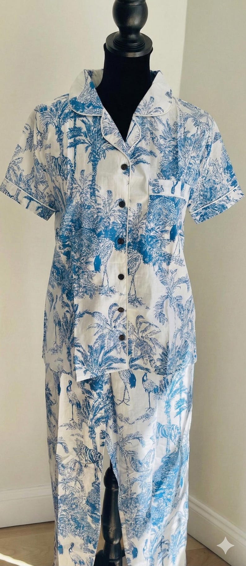 Op de afbeelding: Wit en blauwe pyjamaset met een tropische toile de Jouy-print met palmbomen, zebra's en vogels. De top met korte mouwen heeft een knoopsluiting en een borstzak. De bijpassende broek maakt de set compleet.