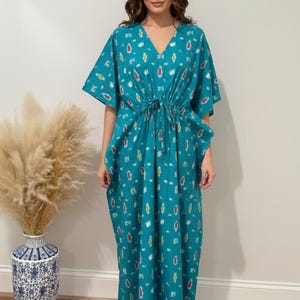 K&ouml;nnte beinhalten: Ein t&uuml;rkisfarbenes Kaftankleid mit V-Ausschnitt, Kordelzug in der Taille und kurzen &Auml;rmeln. Das Kleid hat ein sich wiederholendes Muster aus bunten, abstrakten Formen. Das Model tr&auml;gt das Kleid. Im Hintergrund eine Vase mit getrockneten Pflanzen.