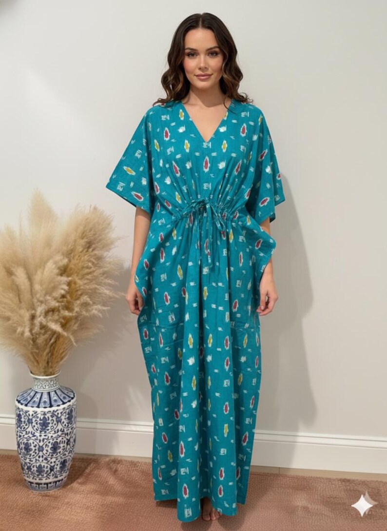 K&ouml;nnte beinhalten: Ein t&uuml;rkisfarbenes Kaftankleid mit V-Ausschnitt, Kordelzug in der Taille und kurzen &Auml;rmeln. Das Kleid hat ein sich wiederholendes Muster aus bunten, abstrakten Formen. Das Model tr&auml;gt das Kleid. Im Hintergrund eine Vase mit getrockneten Pflanzen.