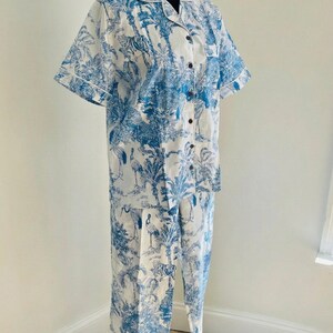 Op de afbeelding: Een tweedelige pyjamaset in wit en blauw. De top met korte mouwen en de broek hebben een toile-de-jouy-print met tijgers, vogels en tropisch gebladerte. De top heeft een knoopsluiting en witte bies.