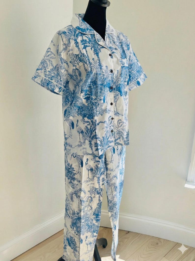Op de afbeelding: Een tweedelige pyjamaset in wit en blauw. De top met korte mouwen en de broek hebben een toile-de-jouy-print met tijgers, vogels en tropisch gebladerte. De top heeft een knoopsluiting en witte bies.