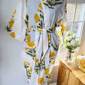 May include: White cotton robe with a yellow floral pattern and green leaves. The robe has a tie waist, pockets, and a shawl collar. The robe is displayed on a black mannequin.