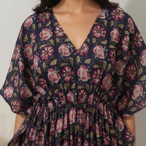K&ouml;nnte beinhalten: Marineblaues Kaftan-Kleid mit V-Ausschnitt und floralem Muster in Rosa und Wei&szlig;. Das Kleid hat kurze &Auml;rmel und eine geraffte Taille. Der Blumendruck enth&auml;lt gr&uuml;ne Bl&auml;tter. Das Kleid ist aus leichtem Stoff gefertigt.