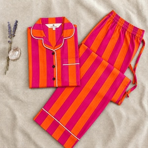 May include: A pajama set featuring a button-down shirt and matching pants with a vibrant vertical stripe pattern in hot pink and bright orange. The shirt has a notched collar, white piping, and a chest pocket. The pants have an elastic waistband.