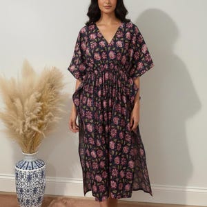 K&ouml;nnte beinhalten: Ein marineblaues Kaftankleid mit Blumendruck, V-Ausschnitt und kurzen &Auml;rmeln. Das Kleid hat eine geraffte Taille und einen Schlitz. Das Blumenmuster enth&auml;lt rosa und gr&uuml;ne Akzente. Das Model tr&auml;gt beige Sandalen.