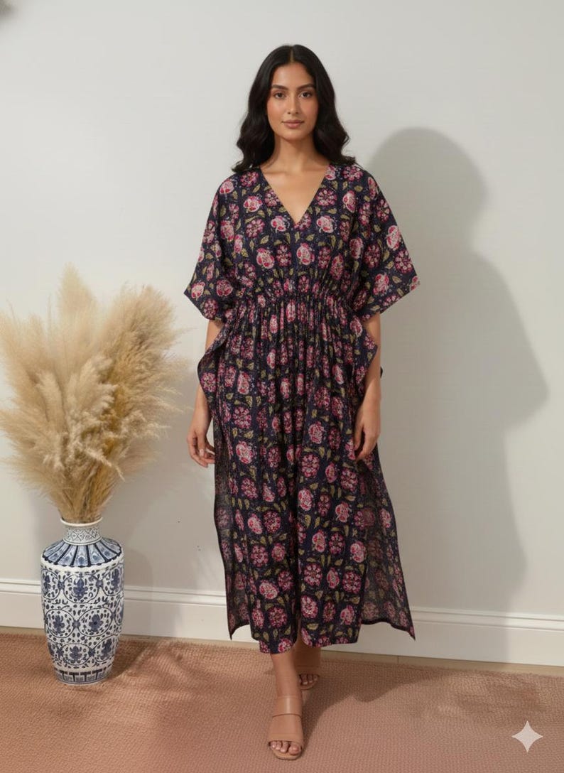 K&ouml;nnte beinhalten: Ein marineblaues Kaftankleid mit Blumendruck, V-Ausschnitt und kurzen &Auml;rmeln. Das Kleid hat eine geraffte Taille und einen Schlitz. Das Blumenmuster enth&auml;lt rosa und gr&uuml;ne Akzente. Das Model tr&auml;gt beige Sandalen.