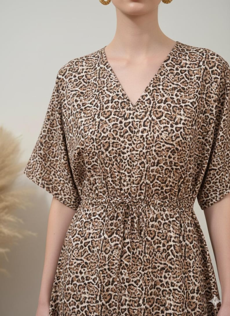 K&ouml;nnte beinhalten: Ein Kleid mit V-Ausschnitt und Leopardenmuster in Braun- und Beiget&ouml;nen. Das Kleid hat kurze &Auml;rmel und eine Kordel in der Taille. Der Stoff wirkt weich und flie&szlig;end, ideal f&uuml;r Freizeitkleidung.