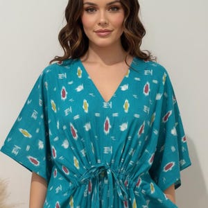 K&ouml;nnte beinhalten: Ein t&uuml;rkisfarbenes Kaftan-Kleid mit V-Ausschnitt und Bindeg&uuml;rtel. Das Kleid hat ein sich wiederholendes Muster aus wei&szlig;en, roten und gelben Formen. Die &Auml;rmel sind weit und der Stoff scheint leicht zu sein. Das Kleid ist ein bequemer, l&auml;ssiger Stil.