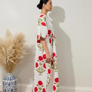 K&ouml;nnte beinhalten: Ein wei&szlig;es, bodenlanges Kaftan mit einem roten Blumenmuster und gr&uuml;nen Bl&auml;ttern. Das Kaftan hat einen V-Ausschnitt und kurze &Auml;rmel. Das Bild zeigt das Kaftan an einer Person. Eine dekorative Vase mit getrockneten Pflanzen befindet sich im Hintergrund.