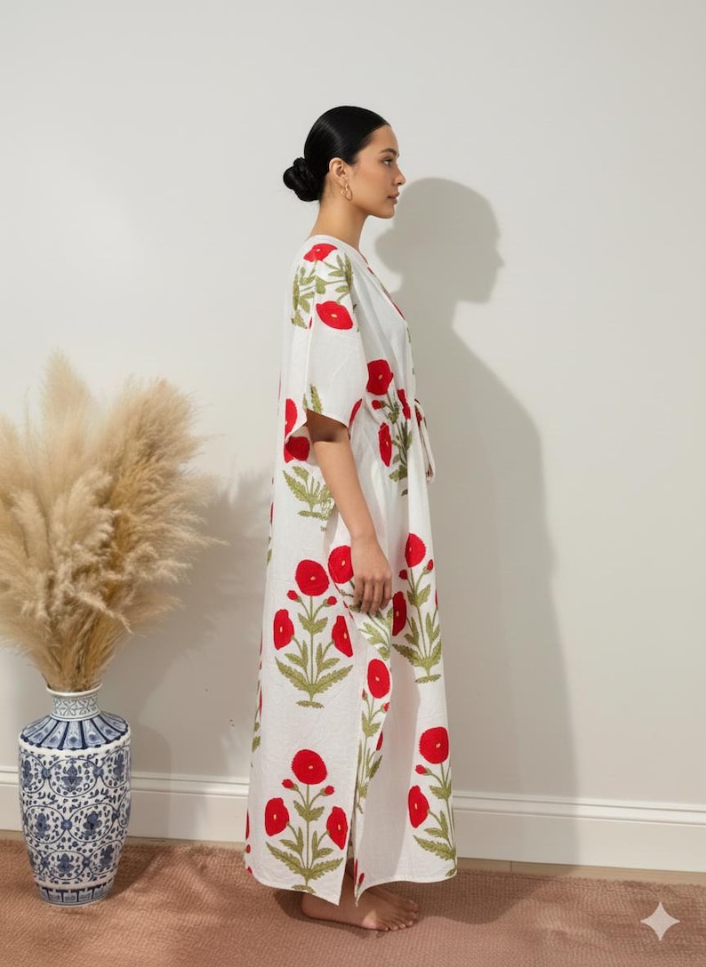 K&ouml;nnte beinhalten: Ein wei&szlig;es, bodenlanges Kaftan mit einem roten Blumenmuster und gr&uuml;nen Bl&auml;ttern. Das Kaftan hat einen V-Ausschnitt und kurze &Auml;rmel. Das Bild zeigt das Kaftan an einer Person. Eine dekorative Vase mit getrockneten Pflanzen befindet sich im Hintergrund.