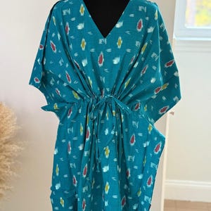 K&ouml;nnte beinhalten: T&uuml;rkisfarbenes Kaftankleid mit V-Ausschnitt und Bindeg&uuml;rtel. Das Kleid hat ein sich wiederholendes Muster aus wei&szlig;en, gelben und roten Formen. Die &Auml;rmel sind weit und flie&szlig;end. Das Kleid wird auf einer schwarzen Schaufensterpuppe pr&auml;sentiert.
