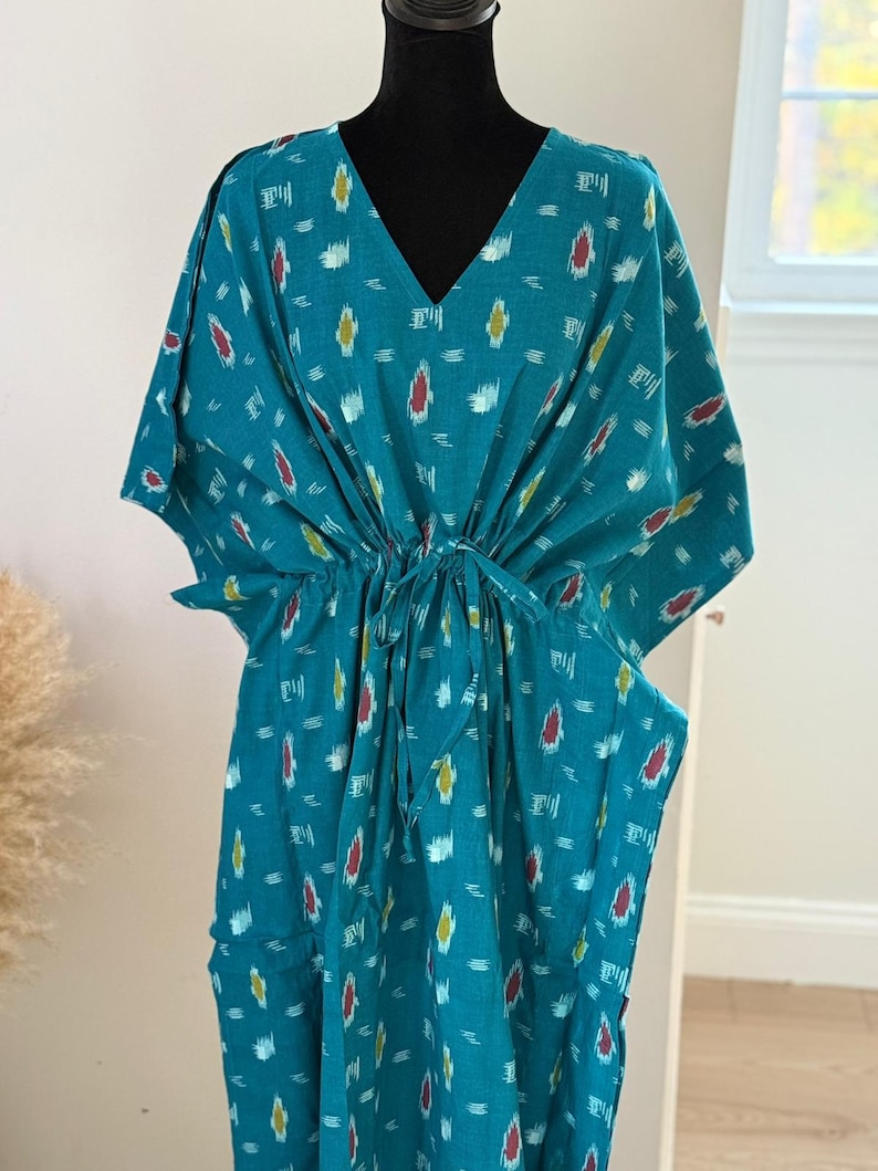 K&ouml;nnte beinhalten: T&uuml;rkisfarbenes Kaftankleid mit V-Ausschnitt und Bindeg&uuml;rtel. Das Kleid hat ein sich wiederholendes Muster aus wei&szlig;en, gelben und roten Formen. Die &Auml;rmel sind weit und flie&szlig;end. Das Kleid wird auf einer schwarzen Schaufensterpuppe pr&auml;sentiert.