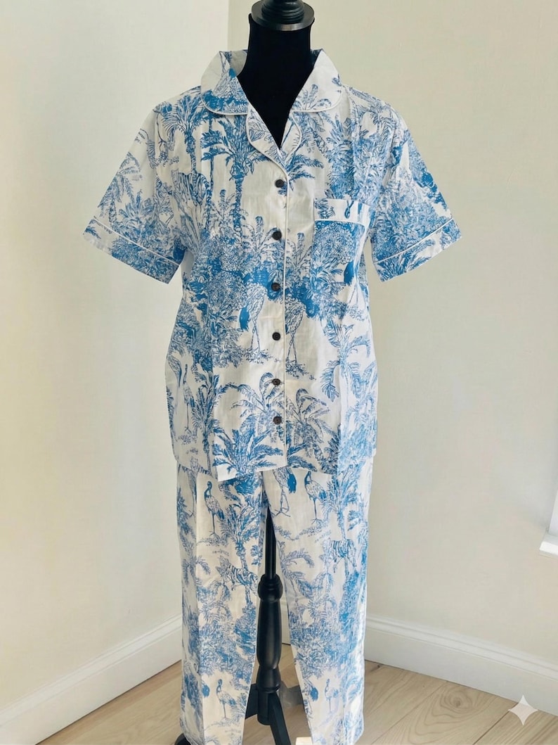 Op de afbeelding: Witte en blauwe pyjamaset met tropische print. De set bestaat uit een shirt met korte mouwen en knopen en een bijpassende broek. De print toont palmbomen, vogels en andere planten. Het shirt heeft een borstzak en witte bies.