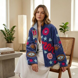 May include: A blue velvet jacket with colorful embroidered patches featuring eyes, stars, a crown, a sun, and a snake. The jacket has a striped collar and a zipper closure. The model is wearing a white dress.