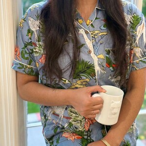 Op de afbeelding: Lichtblauwe pyjamaset met een safari-dierenprint, waaronder zebra's en giraffen. De top met korte mouwen en de broek hebben witte biezen. Een witte mok met een leeuwenkop wordt in de handen gehouden.