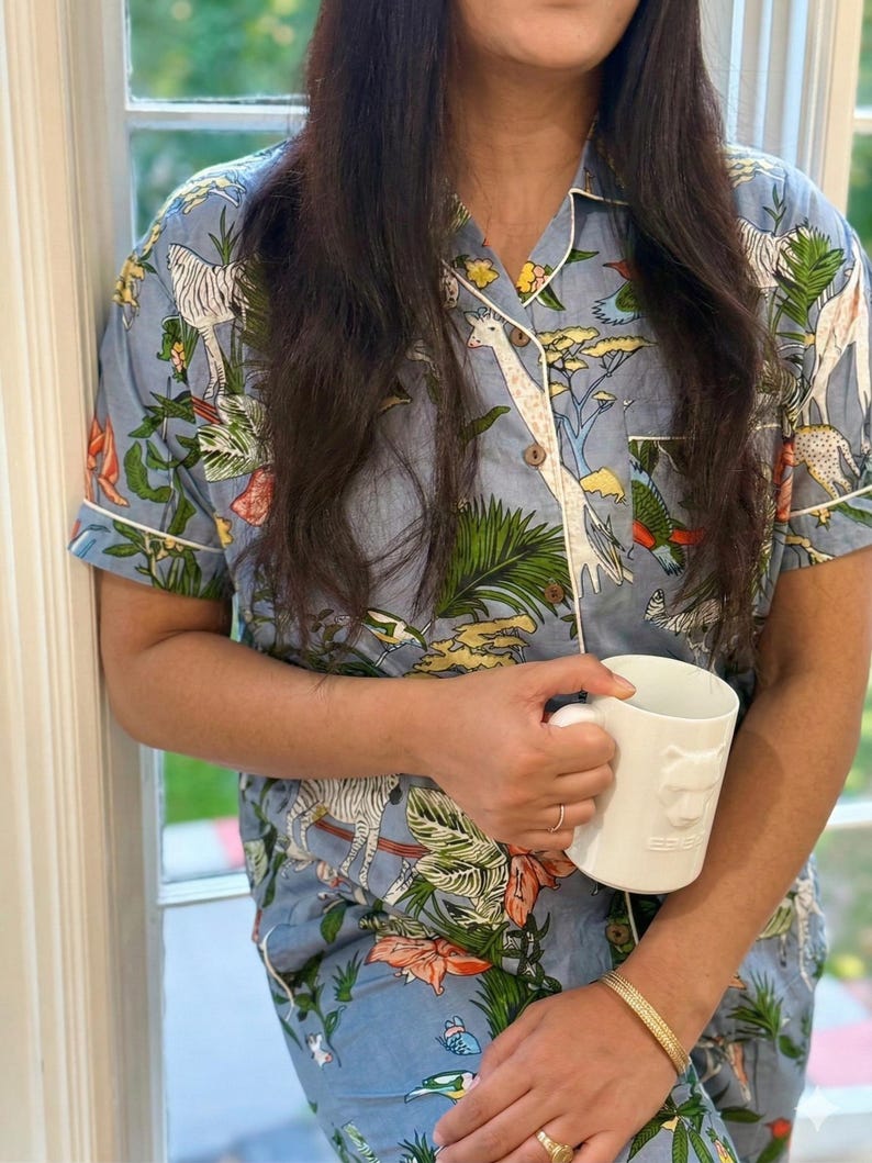 Op de afbeelding: Lichtblauwe pyjamaset met een safari-dierenprint, waaronder zebra's en giraffen. De top met korte mouwen en de broek hebben witte biezen. Een witte mok met een leeuwenkop wordt in de handen gehouden.