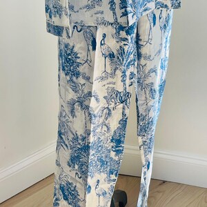 Op de afbeelding: Wit en blauw pyjamaset met een toile de Jouy-patroon met palmbomen, vogels en tijgers. De set bestaat uit een overhemd met knopen en een broek. De stof lijkt licht van gewicht, geschikt voor nachtkleding of loungewear.