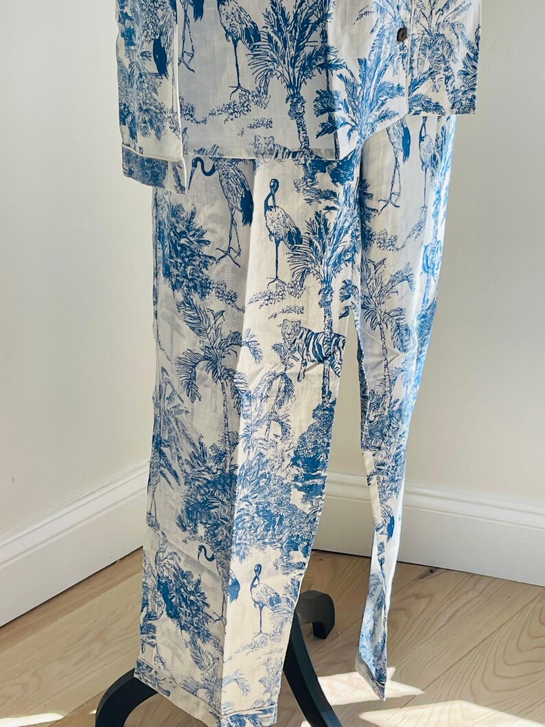 Op de afbeelding: Wit en blauw pyjamaset met een toile de Jouy-patroon met palmbomen, vogels en tijgers. De set bestaat uit een overhemd met knopen en een broek. De stof lijkt licht van gewicht, geschikt voor nachtkleding of loungewear.