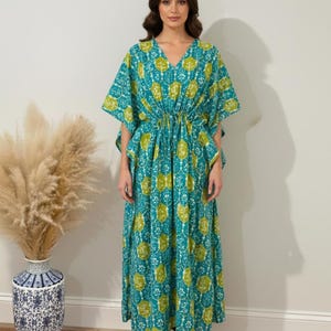 K&ouml;nnte beinhalten: Ein langes, flie&szlig;endes Kaftan-Kleid in T&uuml;rkis und Limettengr&uuml;n. Das Kleid hat einen V-Ausschnitt, kurze &Auml;rmel und eine Kordel in der Taille. Der Stoff ist mit einem sich wiederholenden floralen Muster versehen. Das Model tr&auml;gt das Kleid.