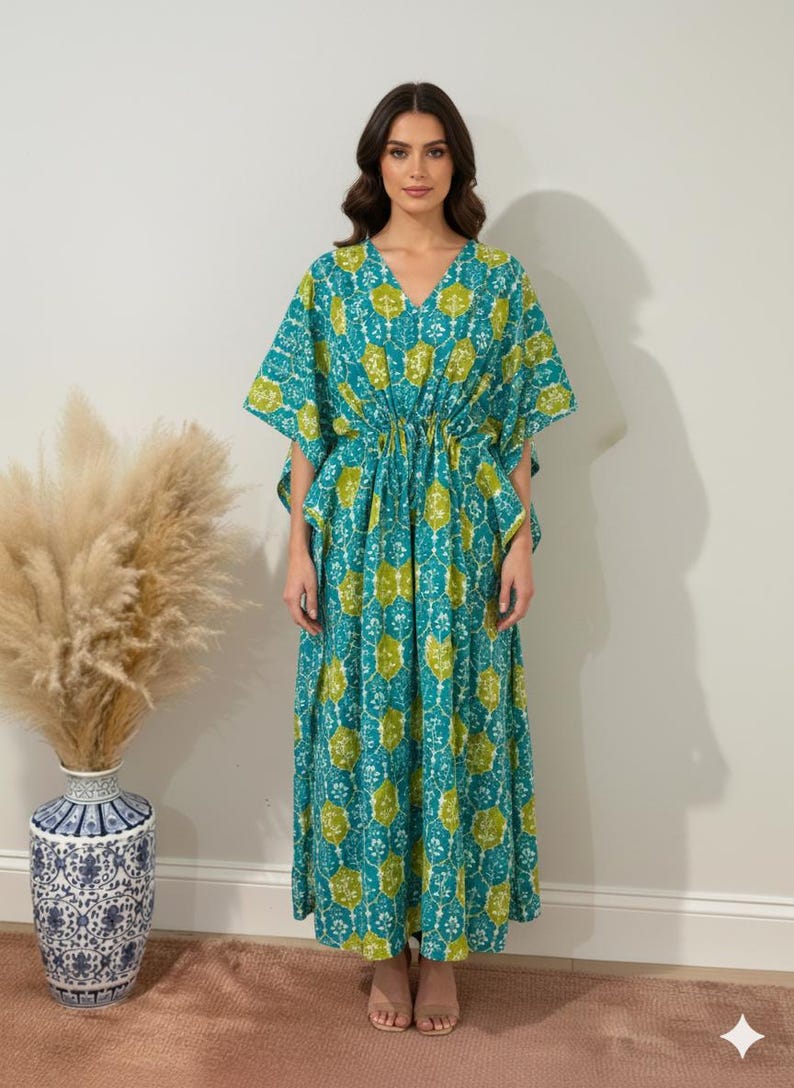 K&ouml;nnte beinhalten: Ein langes, flie&szlig;endes Kaftan-Kleid in T&uuml;rkis und Limettengr&uuml;n. Das Kleid hat einen V-Ausschnitt, kurze &Auml;rmel und eine Kordel in der Taille. Der Stoff ist mit einem sich wiederholenden floralen Muster versehen. Das Model tr&auml;gt das Kleid.