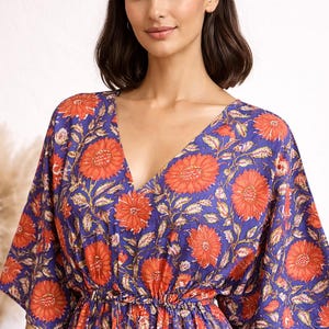 K&ouml;nnte beinhalten: Ein blaues Kleid mit V-Ausschnitt und floralem Muster. Das Kleid hat gro&szlig;e orangefarbene Blumen und gr&uuml;ne Bl&auml;tter auf blauem Grund. Es hat kurze &Auml;rmel und eine geraffte Taille.