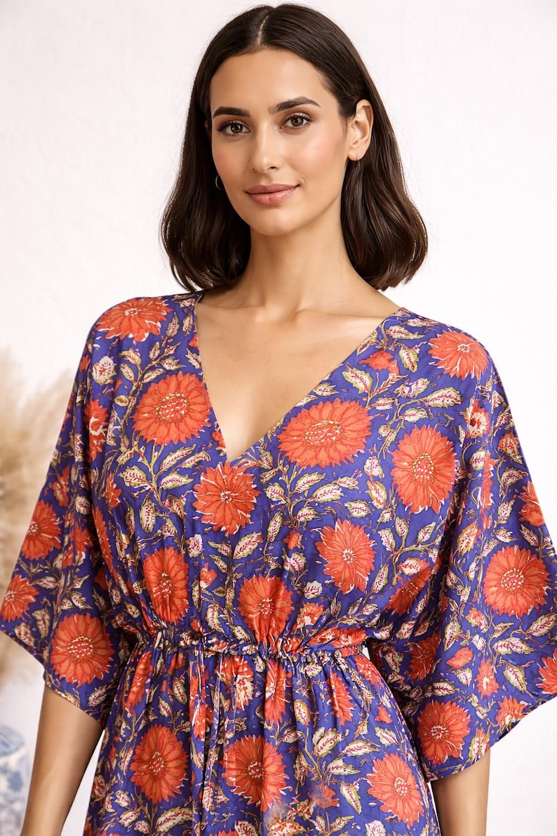 K&ouml;nnte beinhalten: Ein blaues Kleid mit V-Ausschnitt und floralem Muster. Das Kleid hat gro&szlig;e orangefarbene Blumen und gr&uuml;ne Bl&auml;tter auf blauem Grund. Es hat kurze &Auml;rmel und eine geraffte Taille.