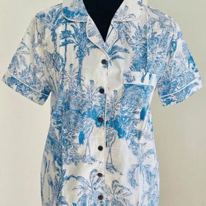 Op de afbeelding: Korte mouwen overhemd met een blauw en wit tropisch patroon. Het overhemd heeft een klassieke kraag, een borstzak en donkerbruine knopen. Het ontwerp bevat palmbomen en andere tropische elementen.