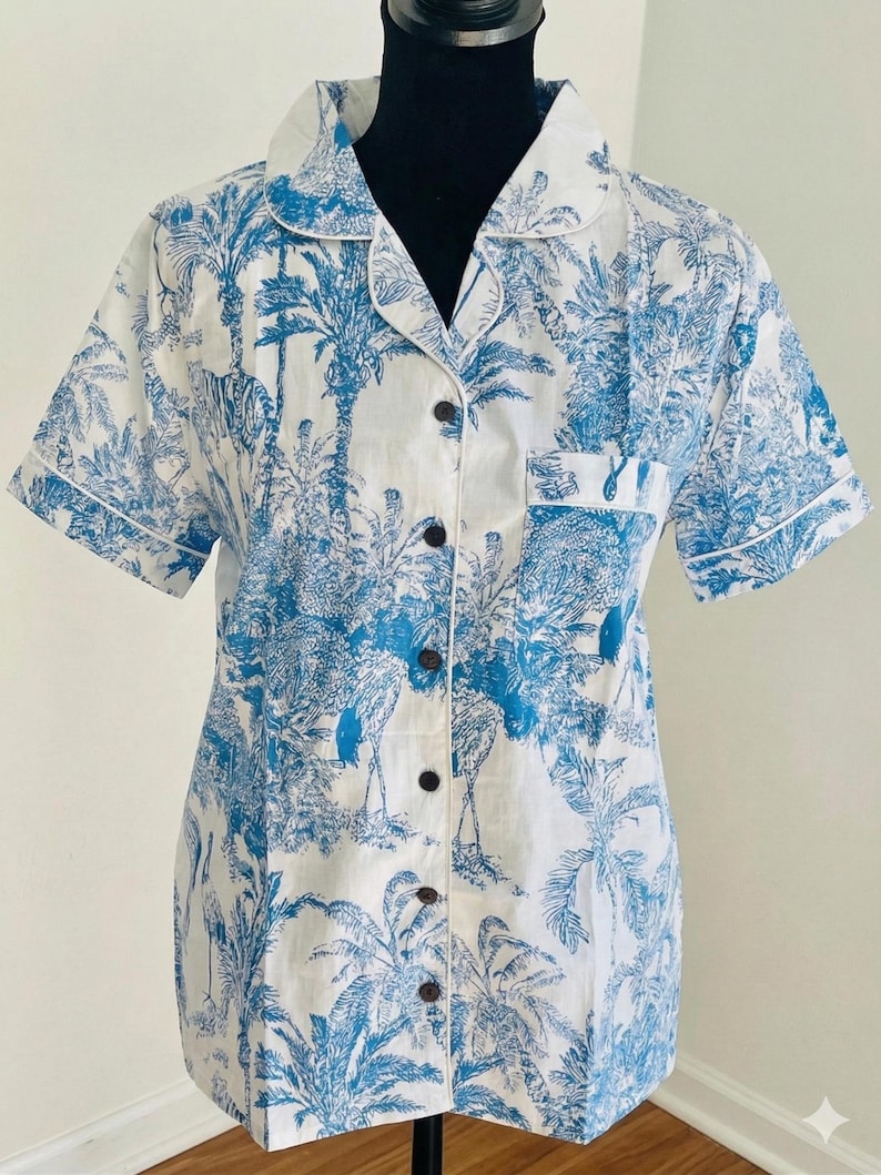 Op de afbeelding: Korte mouwen overhemd met een blauw en wit tropisch patroon. Het overhemd heeft een klassieke kraag, een borstzak en donkerbruine knopen. Het ontwerp bevat palmbomen en andere tropische elementen.