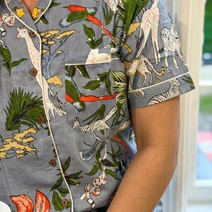 Op de afbeelding: Lichtblauwe pyjama-top met een safari-dierenprint met giraffen, zebra's en vogels. De top met korte mouwen heeft witte biezen, houten knopen en een borstzak. De stof is bedrukt met groene bladeren en oranje bloemen.