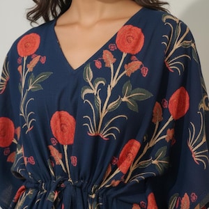 K&ouml;nnte beinhalten: Marineblaues Kaftan-Kleid mit floralem Muster. Das Kleid hat gro&szlig;e rote Blumen mit gr&uuml;nen Bl&auml;ttern und beigen Stielen. Es hat einen V-Ausschnitt und eine Taille mit Kordelzug. Die &Auml;rmel sind kurz und weit.