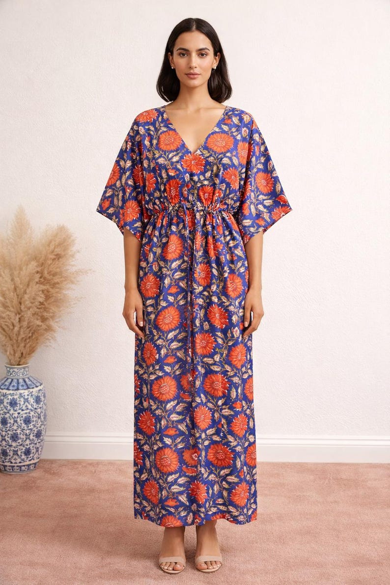 K&ouml;nnte beinhalten: Ein langes, flie&szlig;endes Kleid mit V-Ausschnitt und kurzen &Auml;rmeln. Das Kleid hat ein lebhaftes Blumenmuster in Orange und Wei&szlig; auf tiefblauem Hintergrund. Die Taille ist mit einem Kordelzug gerafft. Das Kleid ist f&uuml;r Freizeitkleidung geeignet.